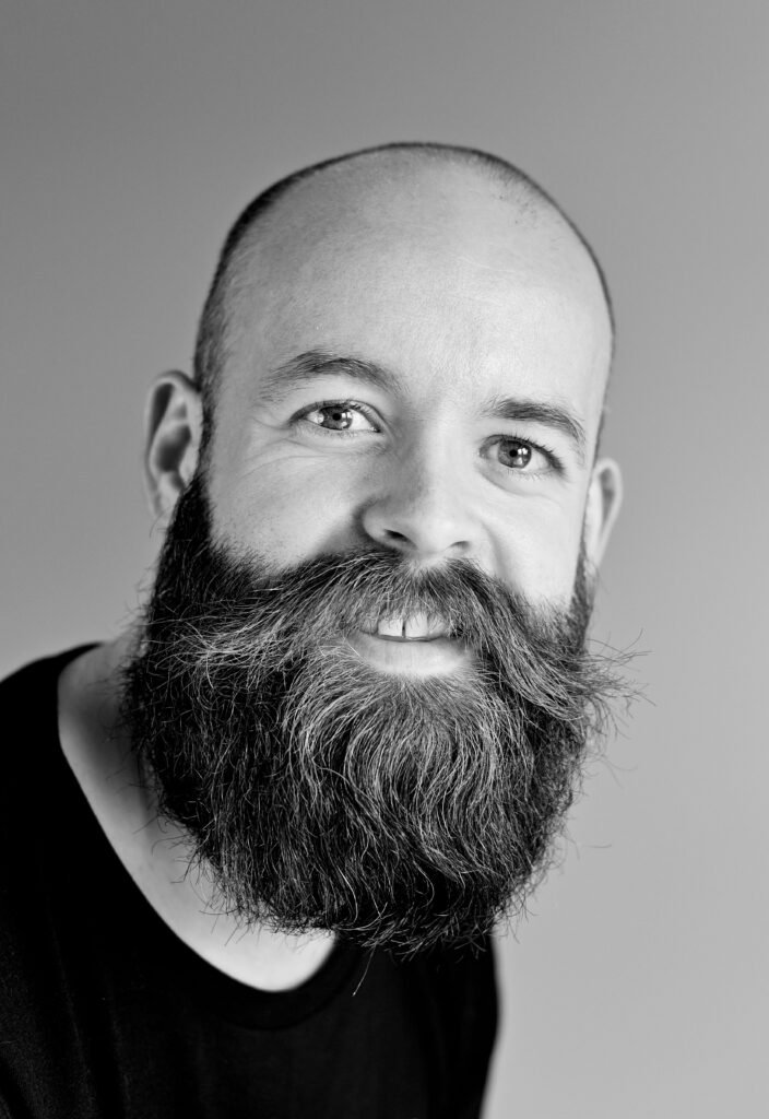 Black and white portrait of a smiling bald man with a large beard in a studio setting.